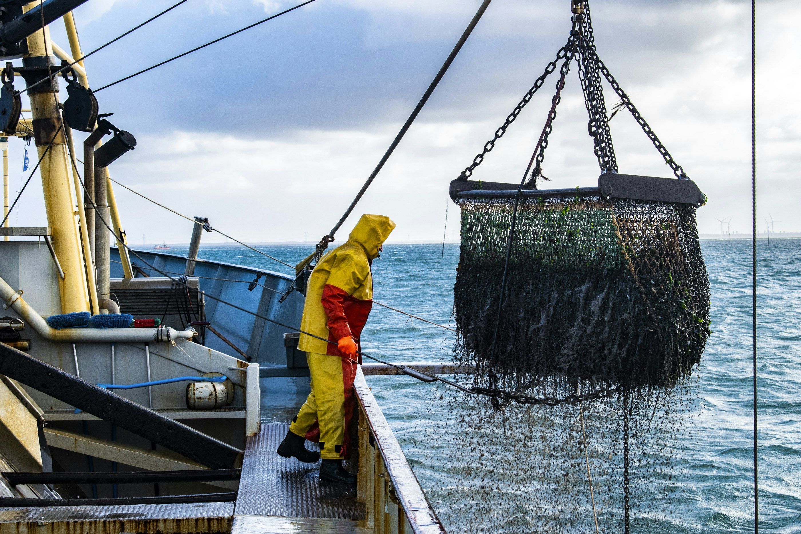 A fisherman in oilskins on deck as a full net is lifted over the side of a fishing vessel